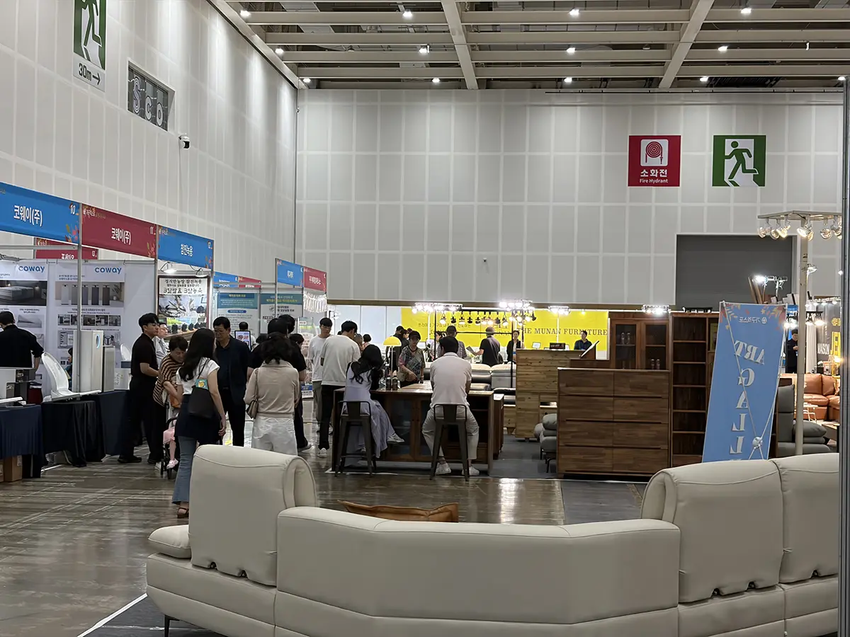 An indoor furniture expo with seating displays in the foreground and multiple vendor booths lined with products and people mingling in the background