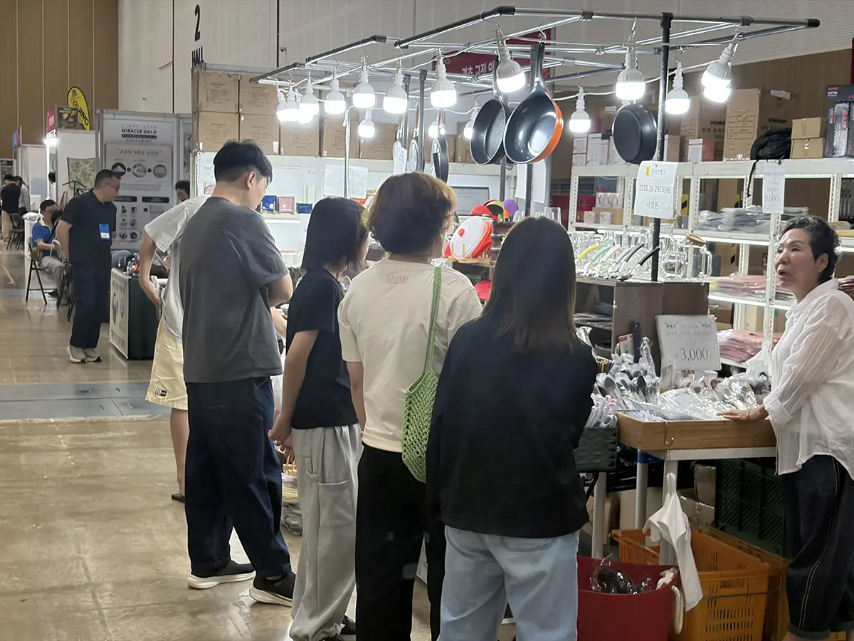 Shoppers at an indoor market stall viewing metal cookware and utensils displayed on shelves under bright hanging lights a vendor stands to the right
