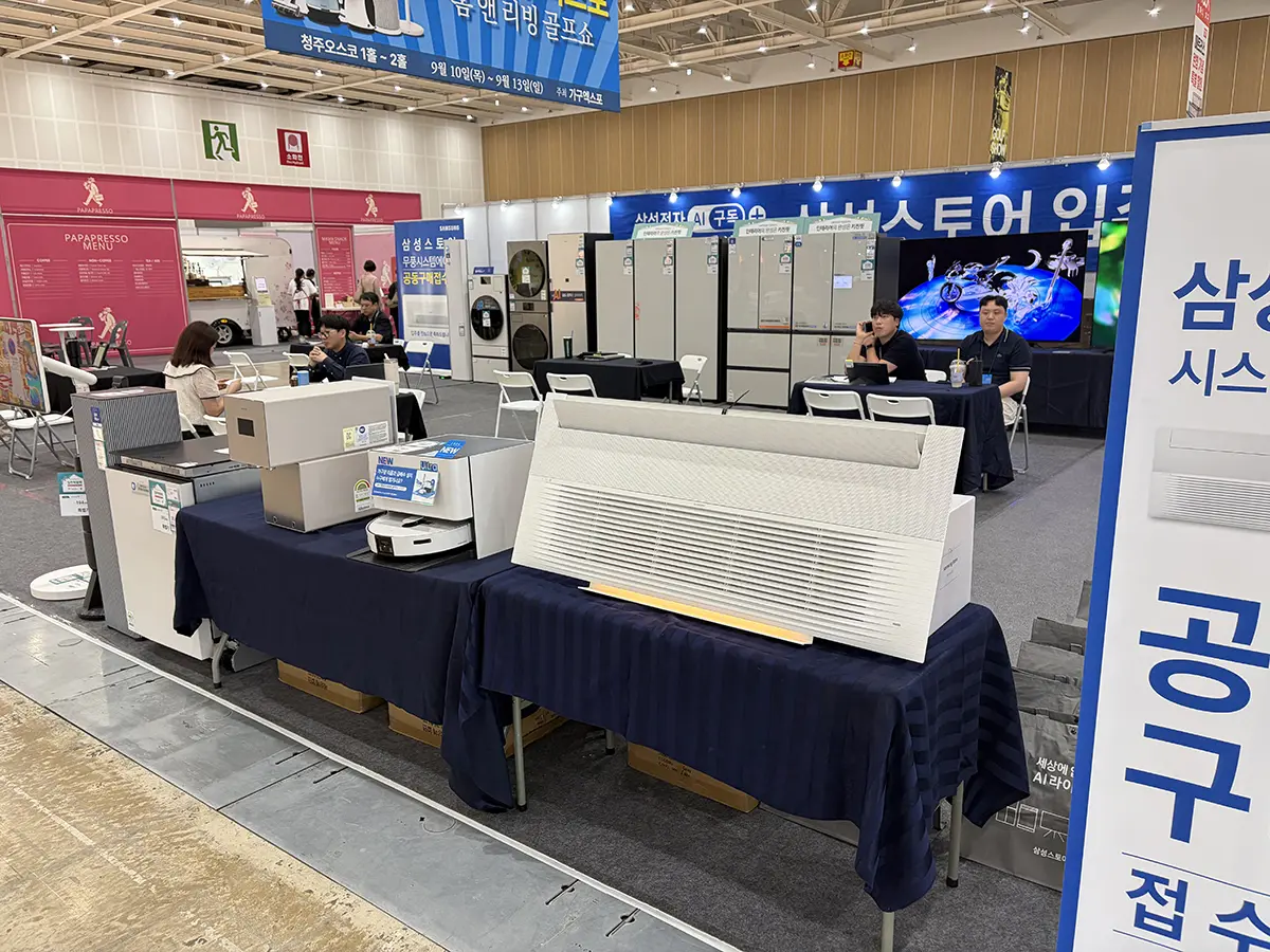 Trade show booth area with large appliances on display refrigerators and related units and attendees seated at tables in the background