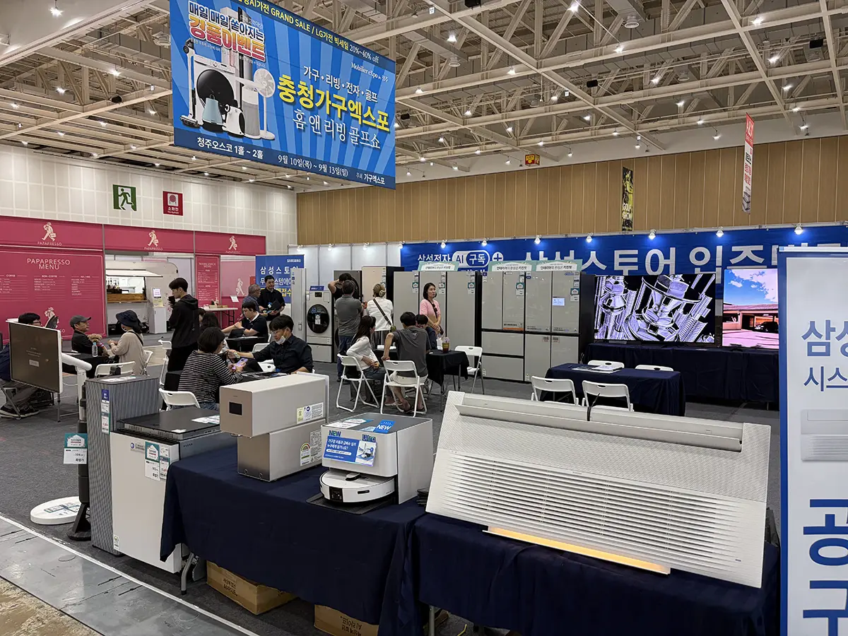 Exhibition hall with people at a home appliance fair displaying large white HVAC components on tables under blue banners