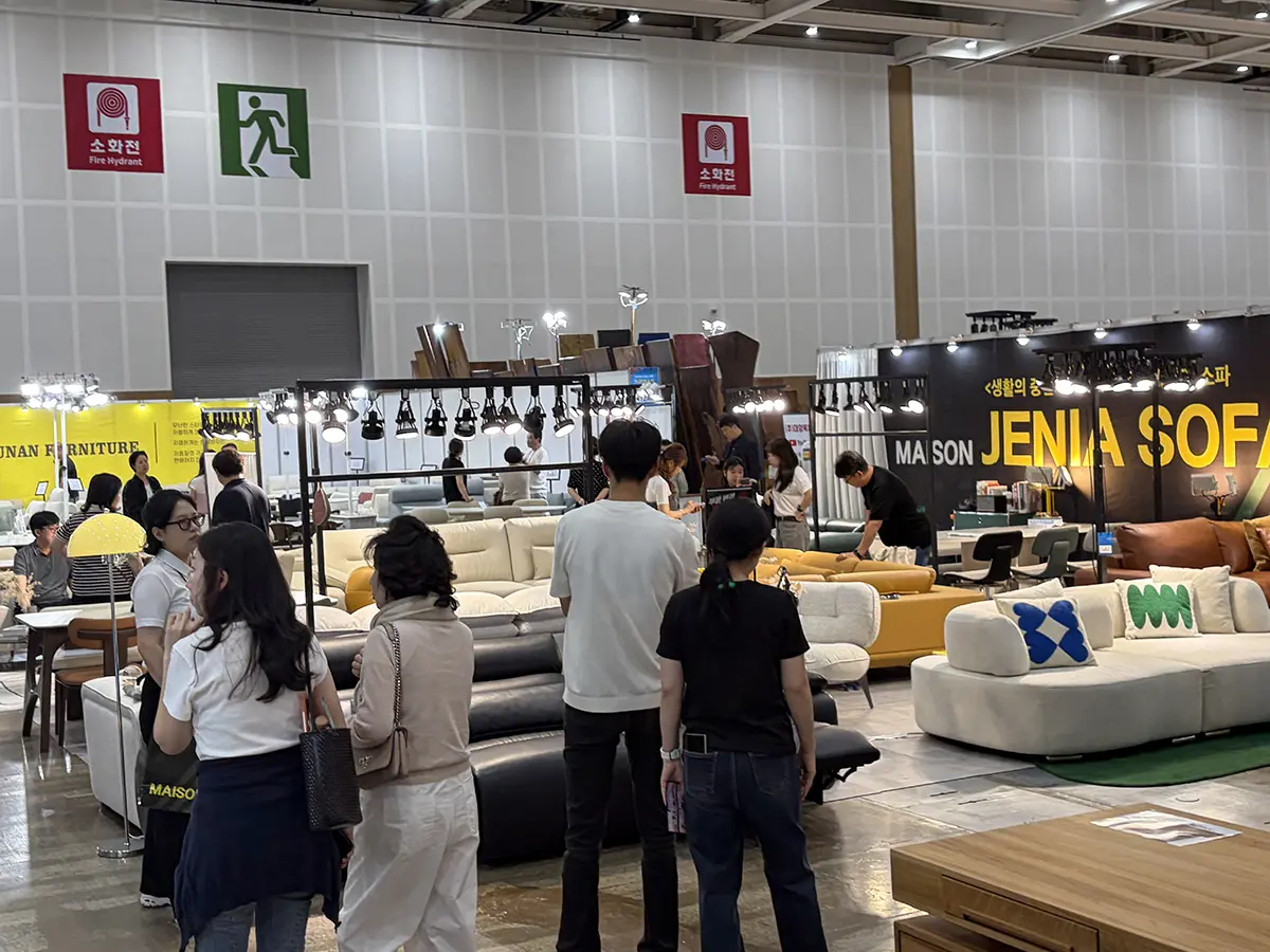 Busy furniture showroom at an exhibition hall with group of people browsing modular sofas and coffee tables under bright display lighting large yellow and black booths in background