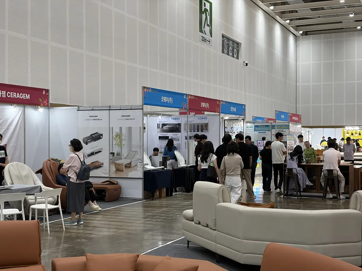 People walk between exhibition booths in a large indoor trade fair with white and brown sofas on display in the foreground