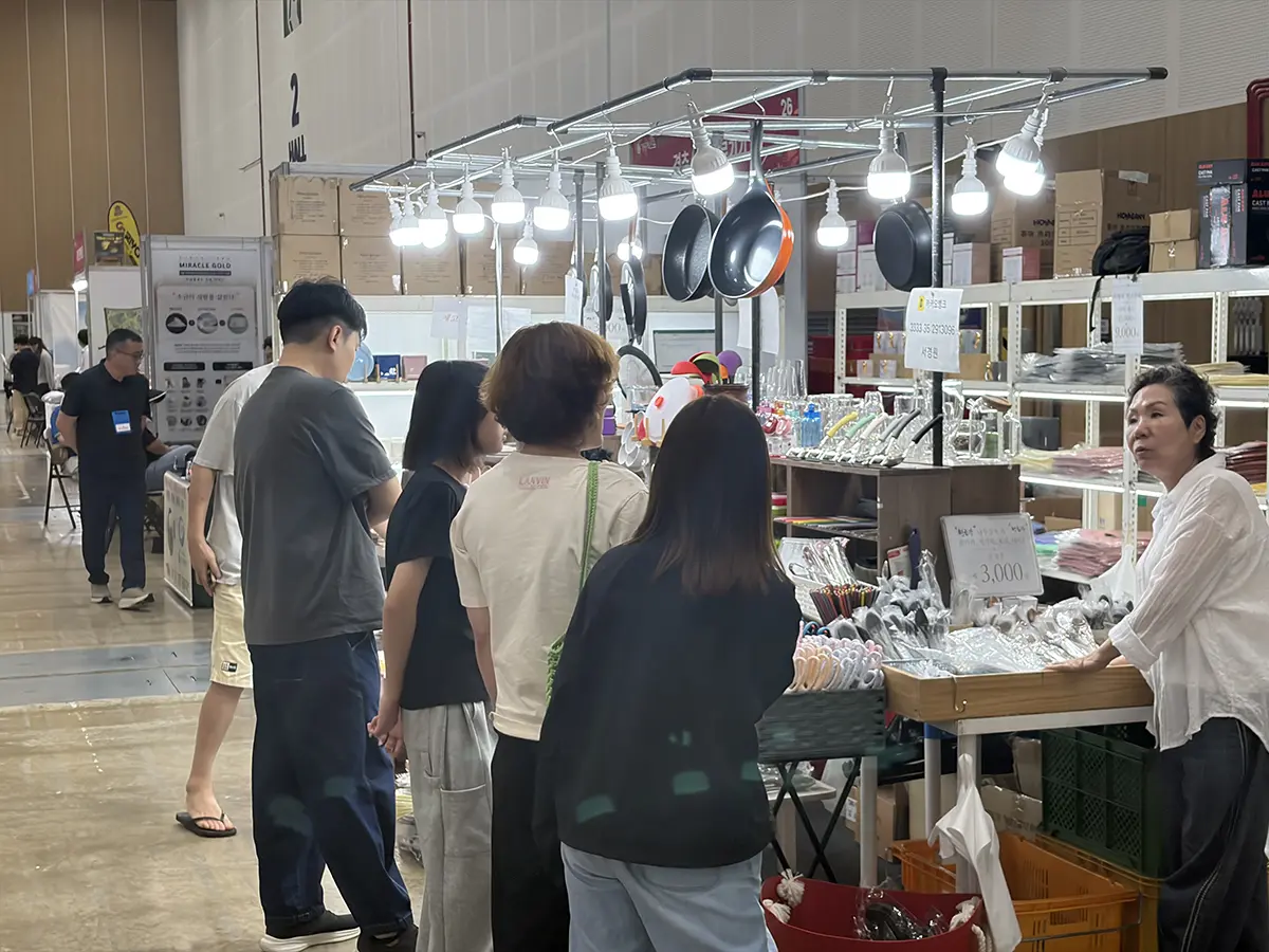 Indoor market stall selling cookware customers browse pans and utensils under bright lights trade show style