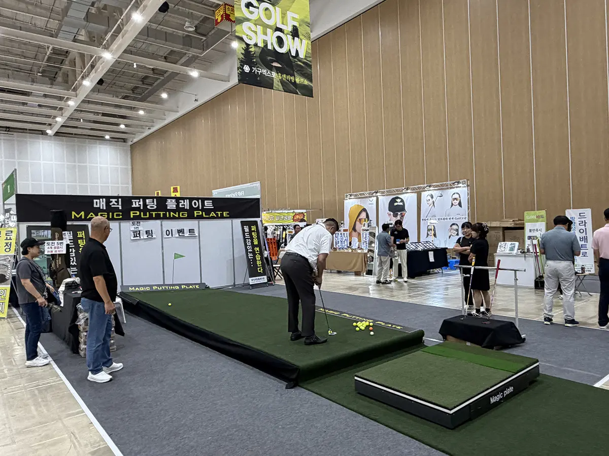 Man in a white shirt and black pants lines up a putt on a green Magic Putting Plate at a trade show demo I view shows onlookers nearby in a large indoor hall