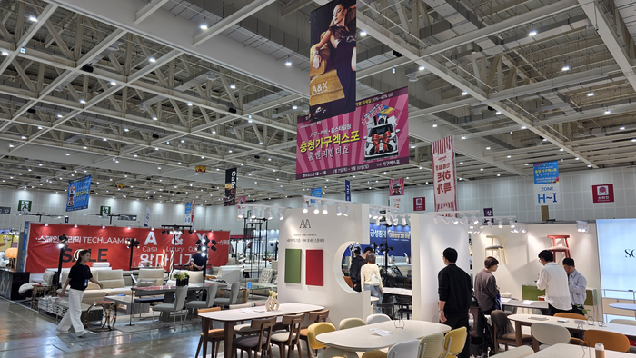 Wide view of a trade show hall with multiple furniture booths hanging banners and visitors browsing displays in bright lighting