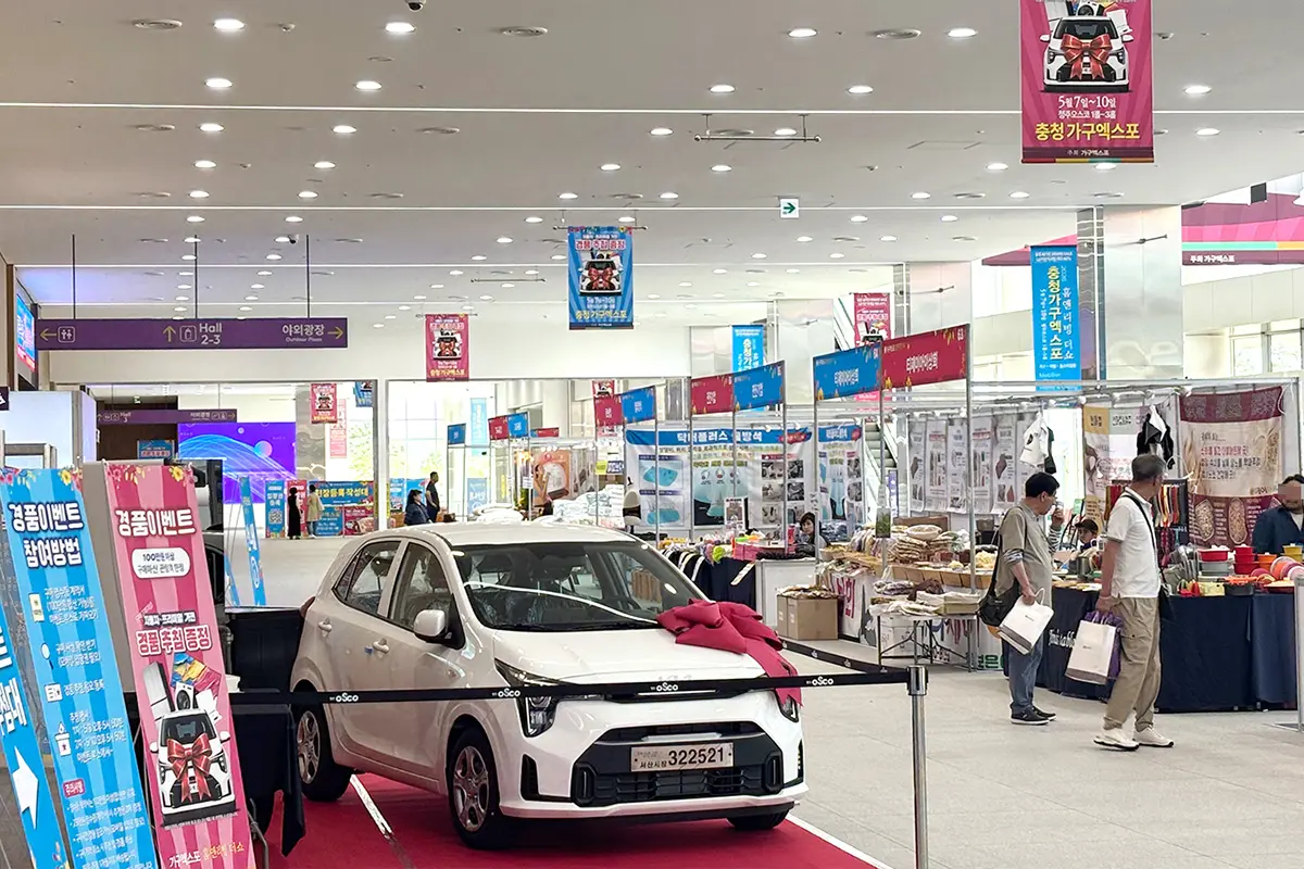 수원가구엑스포전시스케치10 수원 가구엑스포 White compact car on a red carpet display at an indoor expo with a pink bow on the hood and stalls around it