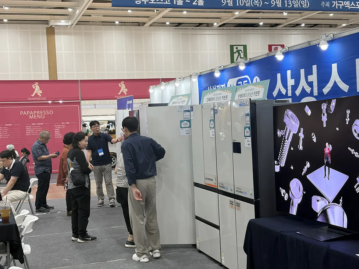 Group of attendees discusses white kitchen appliances at a trade show booth with a pink Pap Espresso sign in the background