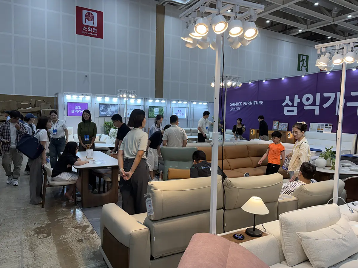 Indoor furniture expo scene with beige and pastel sofas tables and lamps on display as visitors browse the showroom beneath bright overhead lights