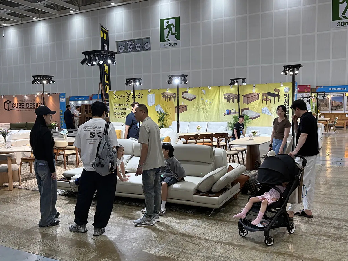 Indoor furniture showroom at a expo with people browsing a white leather sofa set and other wooden furniture displays under bright overhead lights