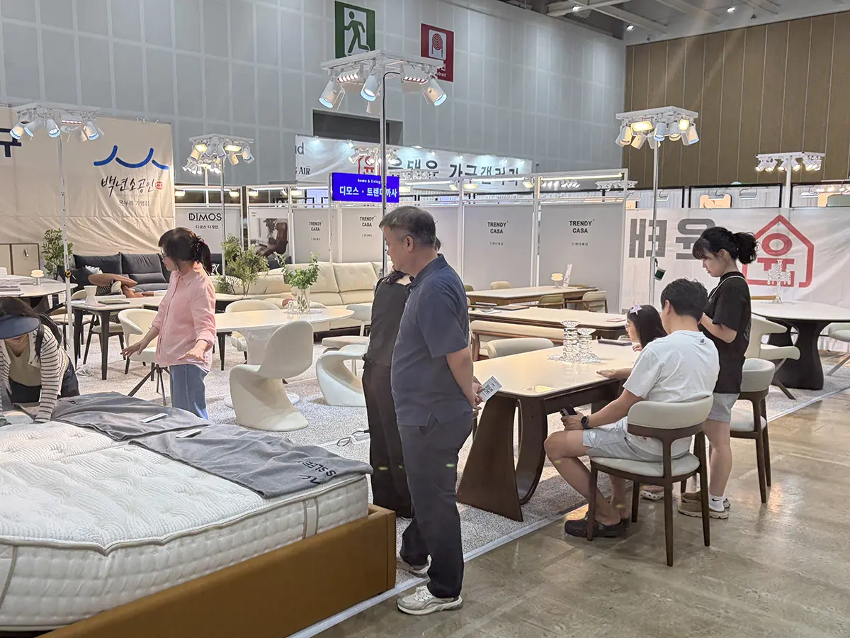 Shoppers examine a large bed with a mattress at a furniture expo surrounded by tables chairs and display areas